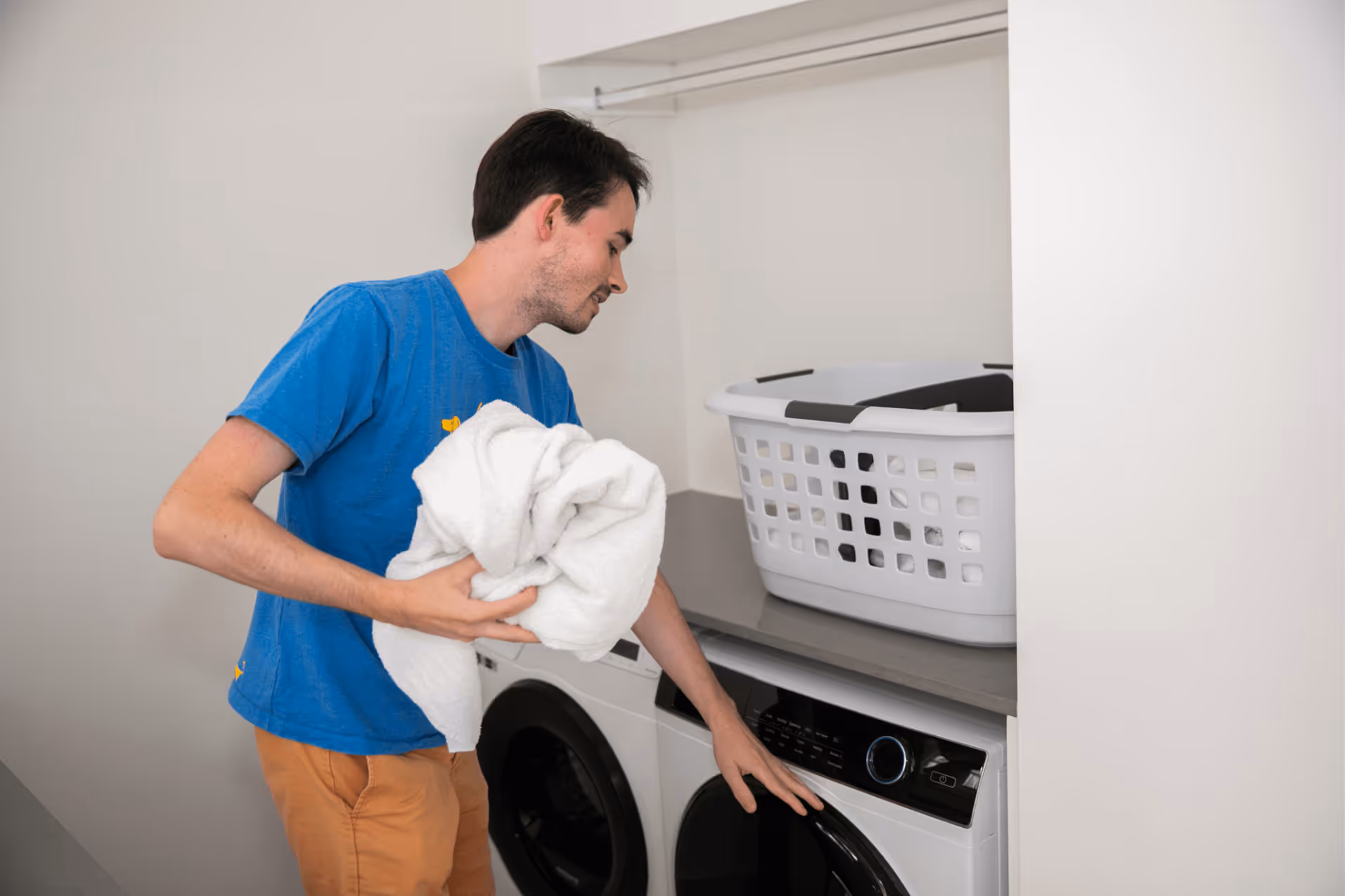 A young man with autism holds a white towel and bends down in front of a washing machine and dryer.
