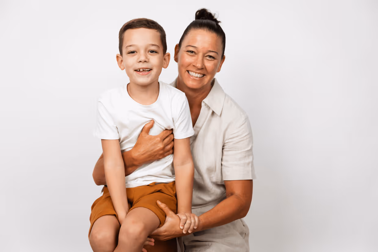 A boy with autism with dark brown hair sits on his mum's lap. They are both wearing white and smiling.