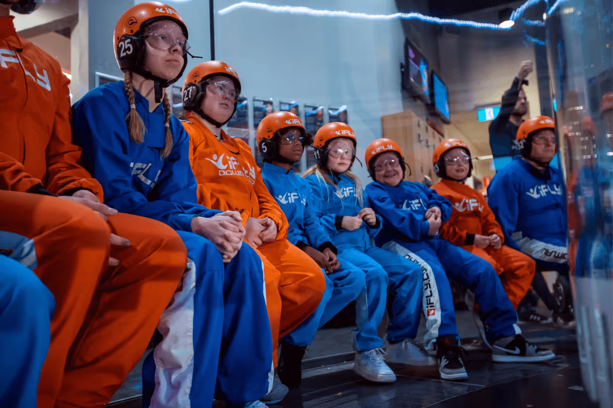 A group of people with disabilities in blue and orange jumpsuits with orange helmets sitting and staring at a glass tube. One smiles at the camera.