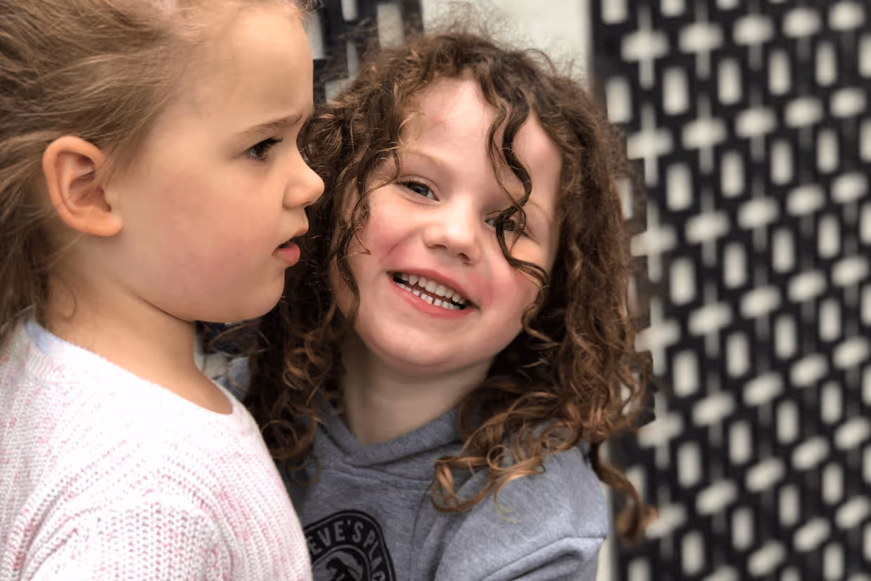 A girl with brown curly hair smiling at the camera and hugging her sister, who does not look at the camera.