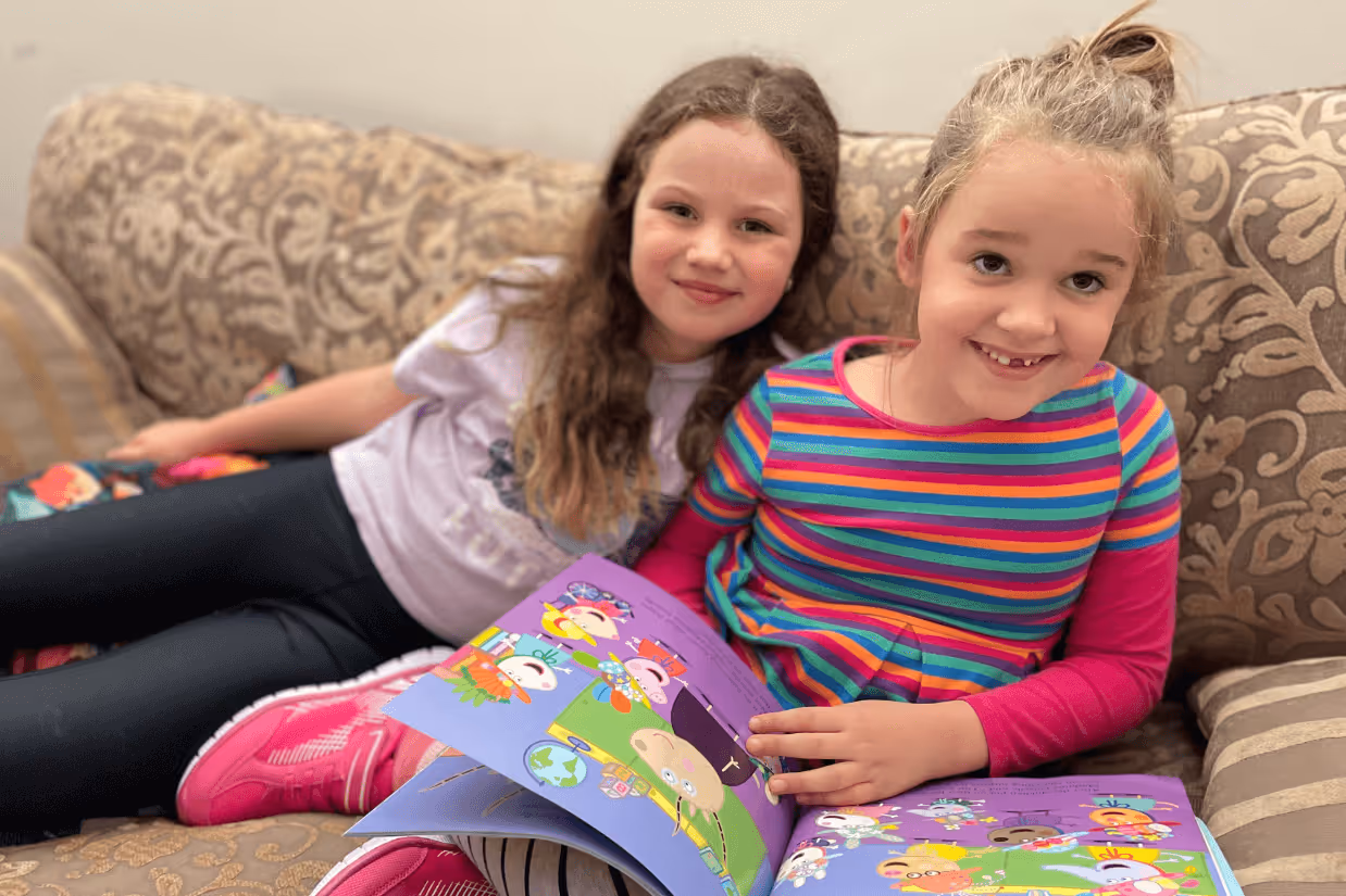Two girls sitting on a brown couch. One is holding a book open. They are both smiling at the camera.