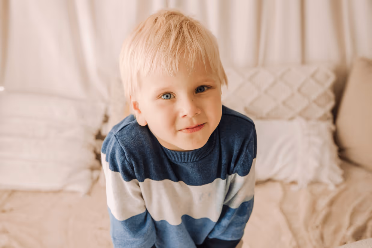 An autistic boy with light blonde hair, blue eyes and a blue striped jumper smiles with closed lips.