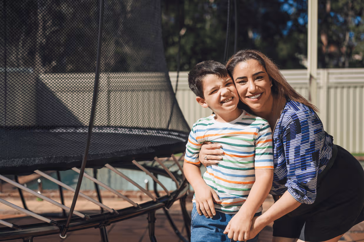 An autistic boy with a striped shirt and denim shirt and his mum hug in front of a Springfree trampoline.