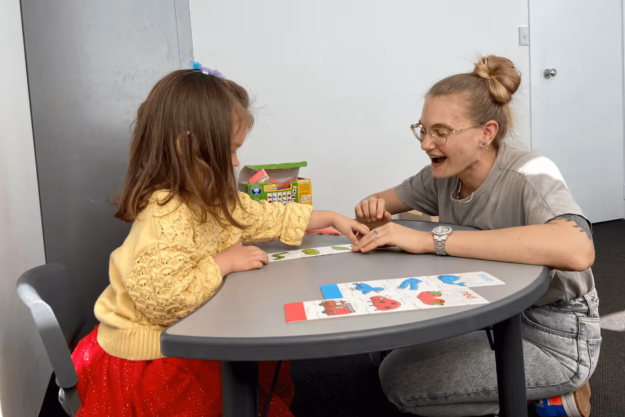 A woman with blonde hair, a grey t-shirt and glasses looks at picture cards with a young girl wearing a yellow jumper and red skirt.