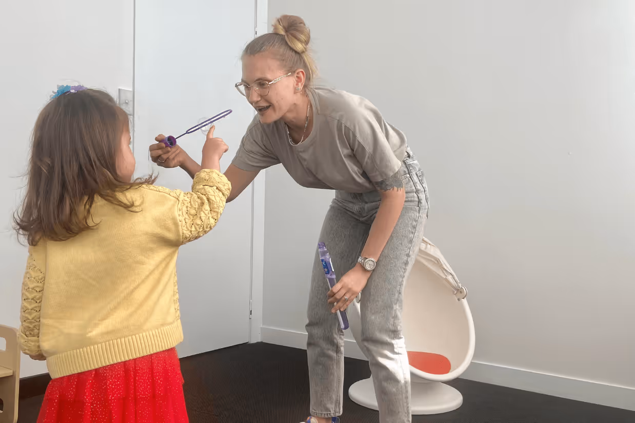 A woman with blonde hair a grey shirt and jeans blows bubbles with a young girl wearing a yellow shirt and red skirt.