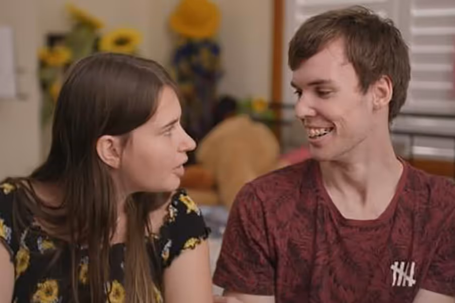 A woman with dark brown hair and a black shirt with yellow sunflowers and a young man with short brown hair, braces and a maroon shirt smile at each other. They are both on the autism spectrum.t