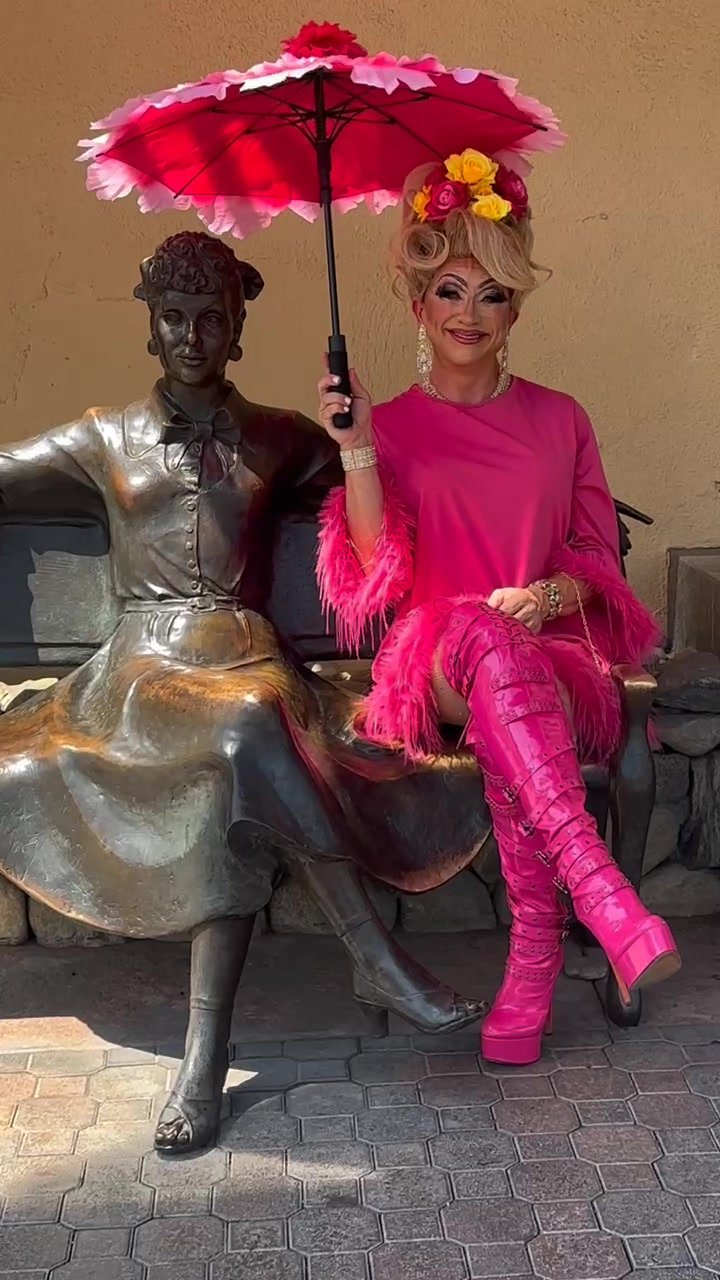 Drag queen with a pink parasol seated beside the Lucille Ball bench statue in Palm Springs