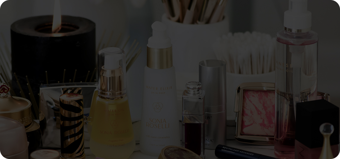 Collection of skincare and makeup products including bottles, a lipstick, a compact with pink blush, and a lit black candle on a white surface.