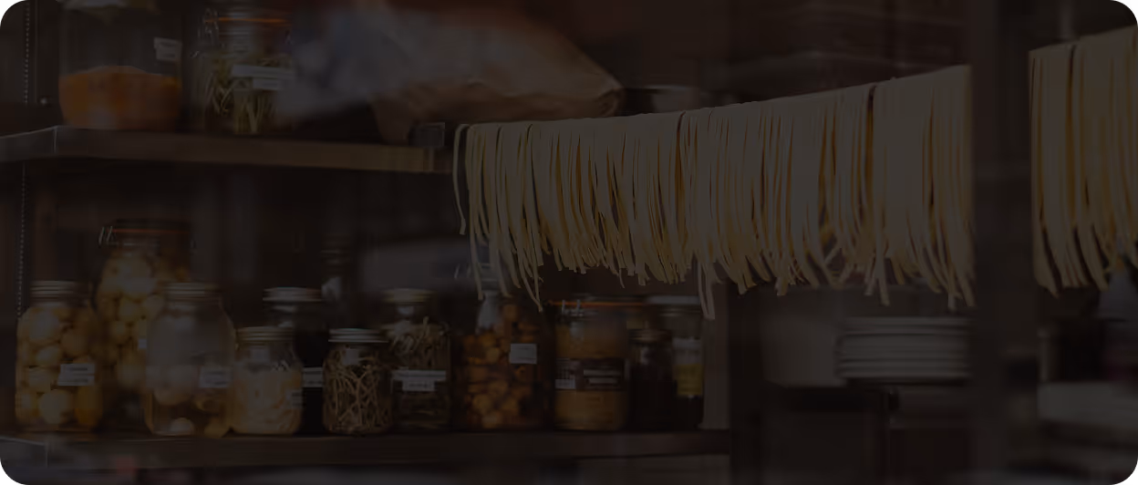 Fresh pasta strands hanging to dry in a kitchen with jars of preserved food on shelves in the background.