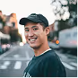 Young man wearing a black cap and dark shirt smiling outdoors in an urban setting.