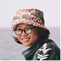 Smiling person wearing glasses and a floral bucket hat, with a blurred outdoor background.