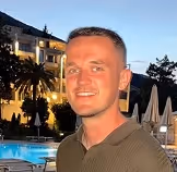 Smiling man with short hair standing in front of a swimming pool and building at dusk.