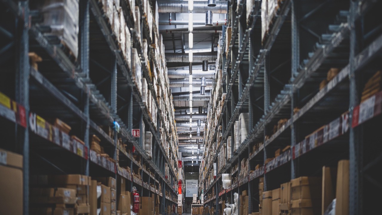 A long warehouse aisle with tall metal racks full of boxes and wrapped stock on both sides