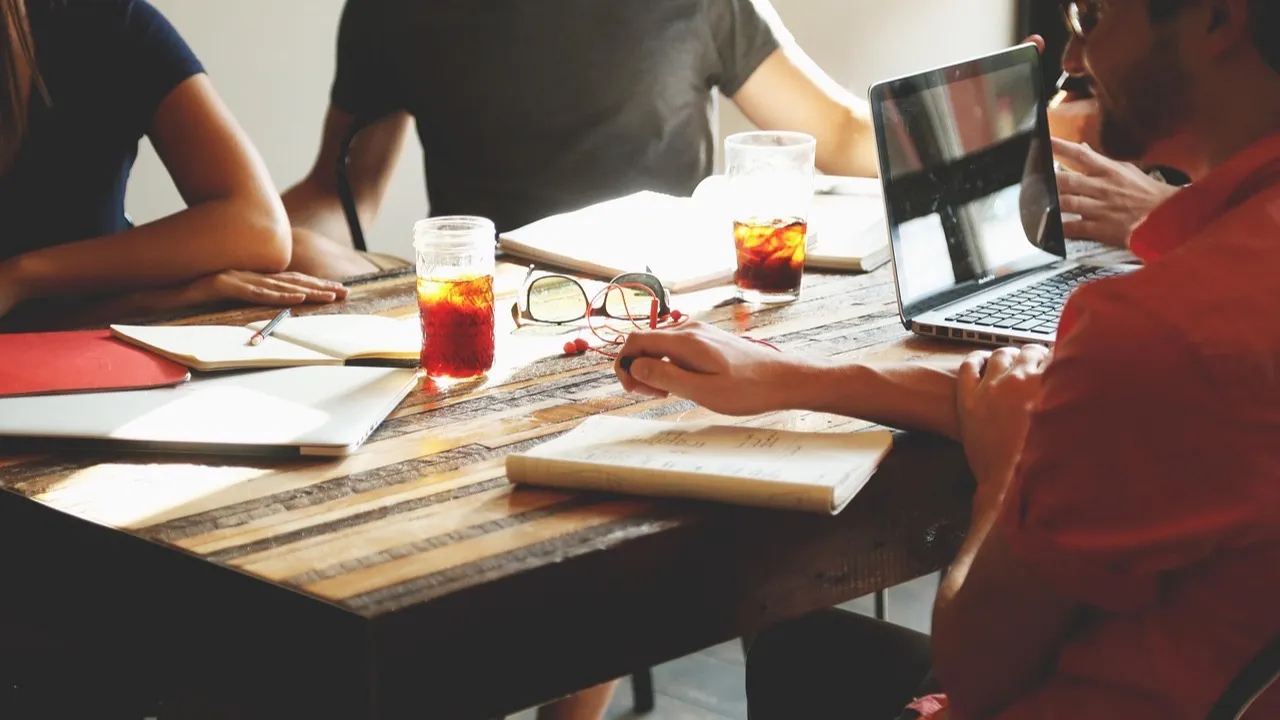A small team sits around a wooden table with laptops and notebooks, planning their work together.