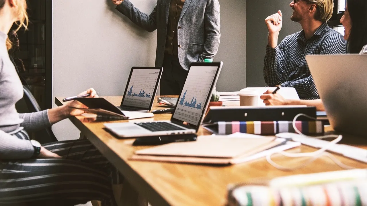 A team meets around a table with laptops while one person points to the wall.