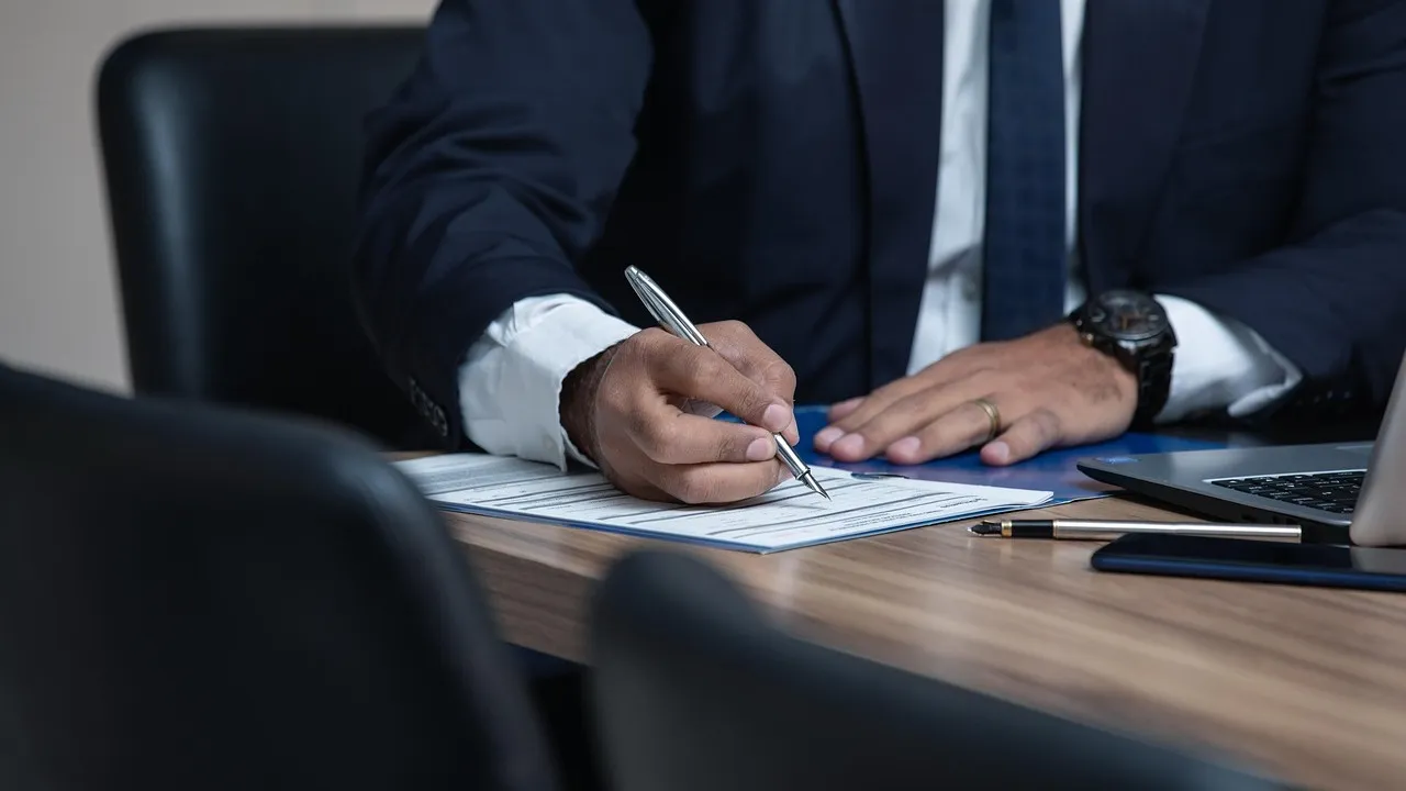 A man in a suit signs a form at a desk, with a laptop and pens beside him.