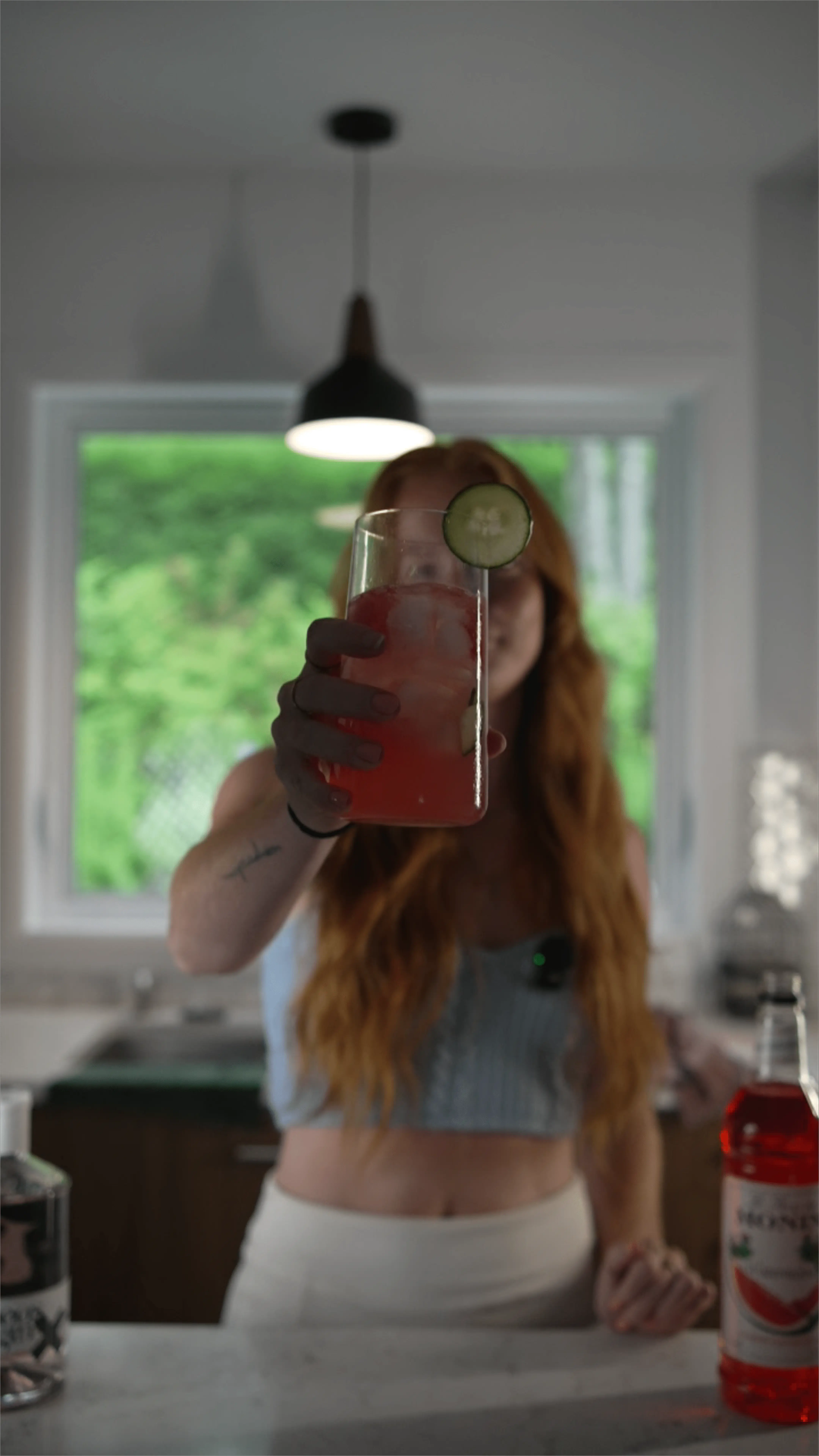 Person with long red hair holding a glass of pink cocktail with ice and cucumber slice towards the camera in a kitchen.