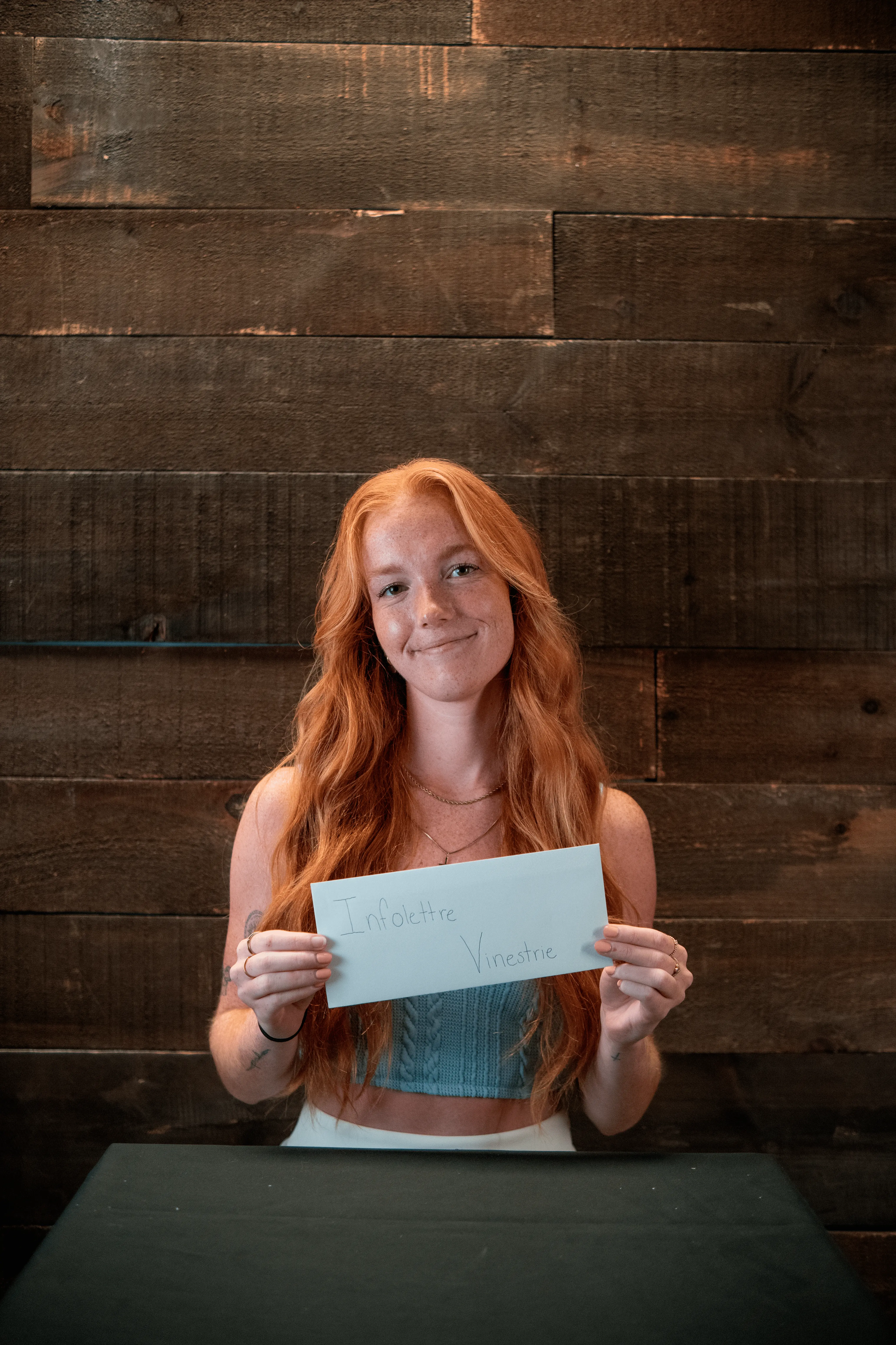 Young woman with long red hair holding an envelope with ‘Infolettre Vinestrie’ handwritten on it in front of a wooden wall.