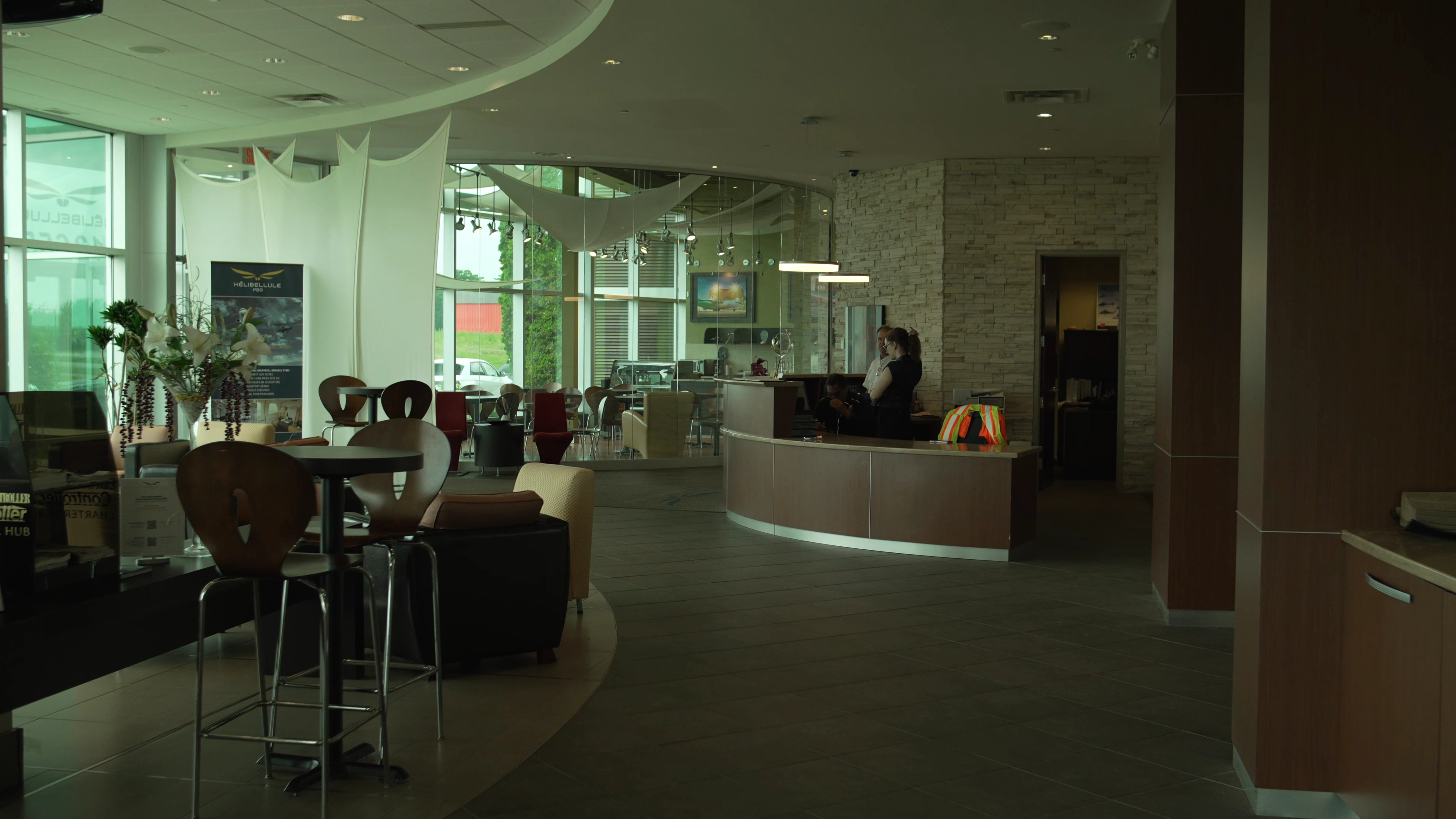 Modern office lobby with curved reception desk, two people standing behind it, and seating area with tables and chairs.