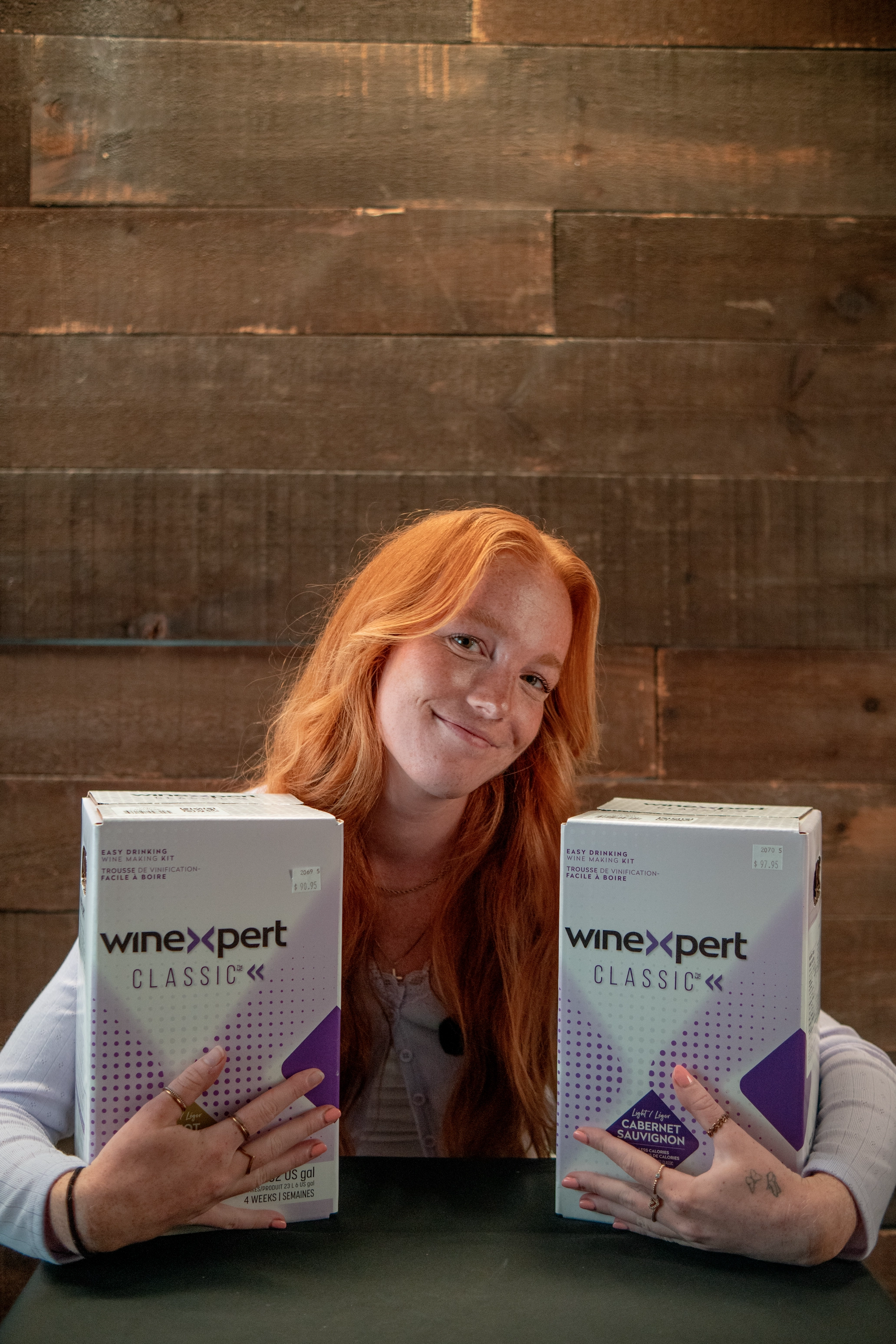 Smiling red-haired woman sitting at a table holding two WineXpert Classic wine making kits.
