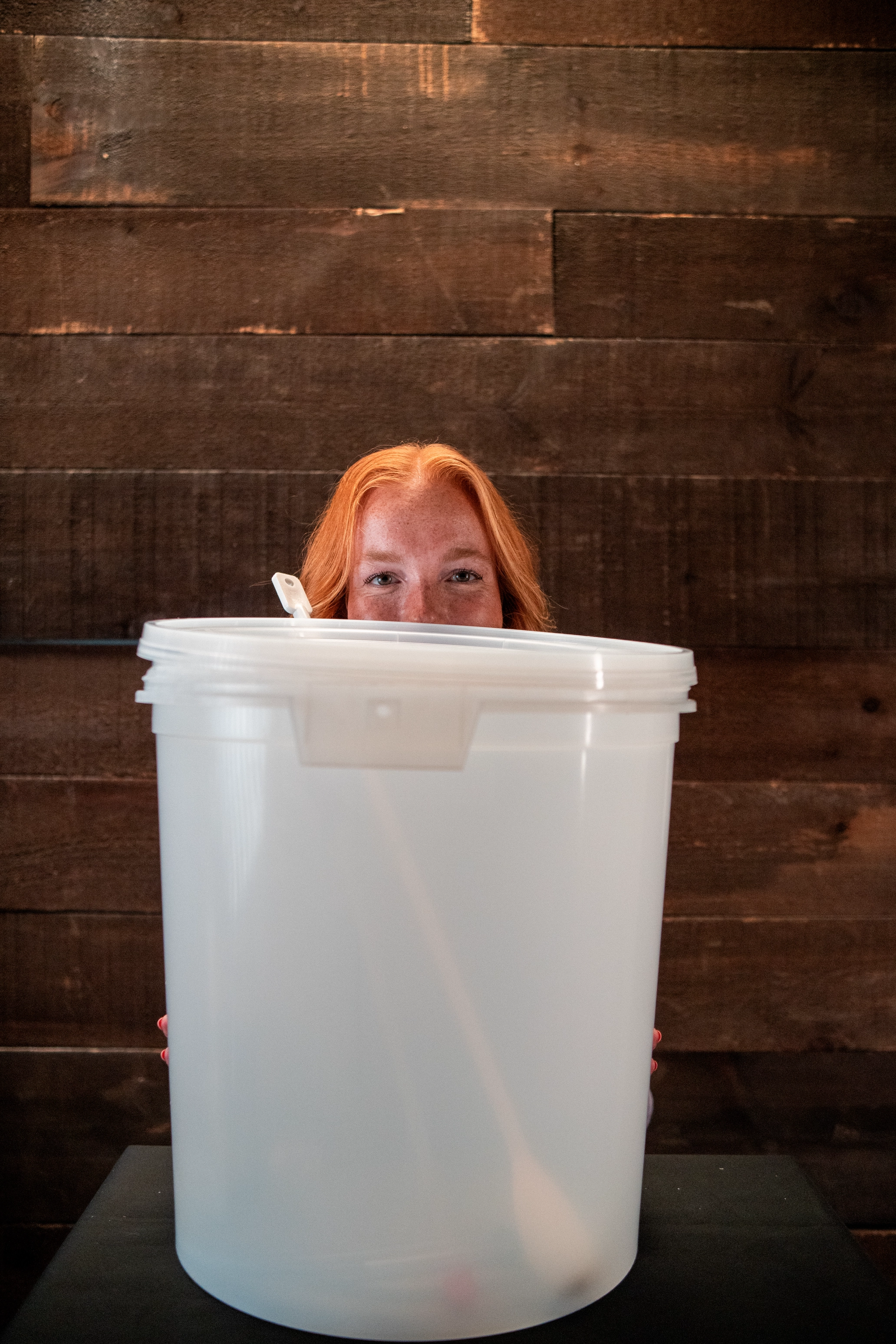 Red-haired person peeking over a large white plastic bucket with a wooden spoon inside, against a dark wooden wall.