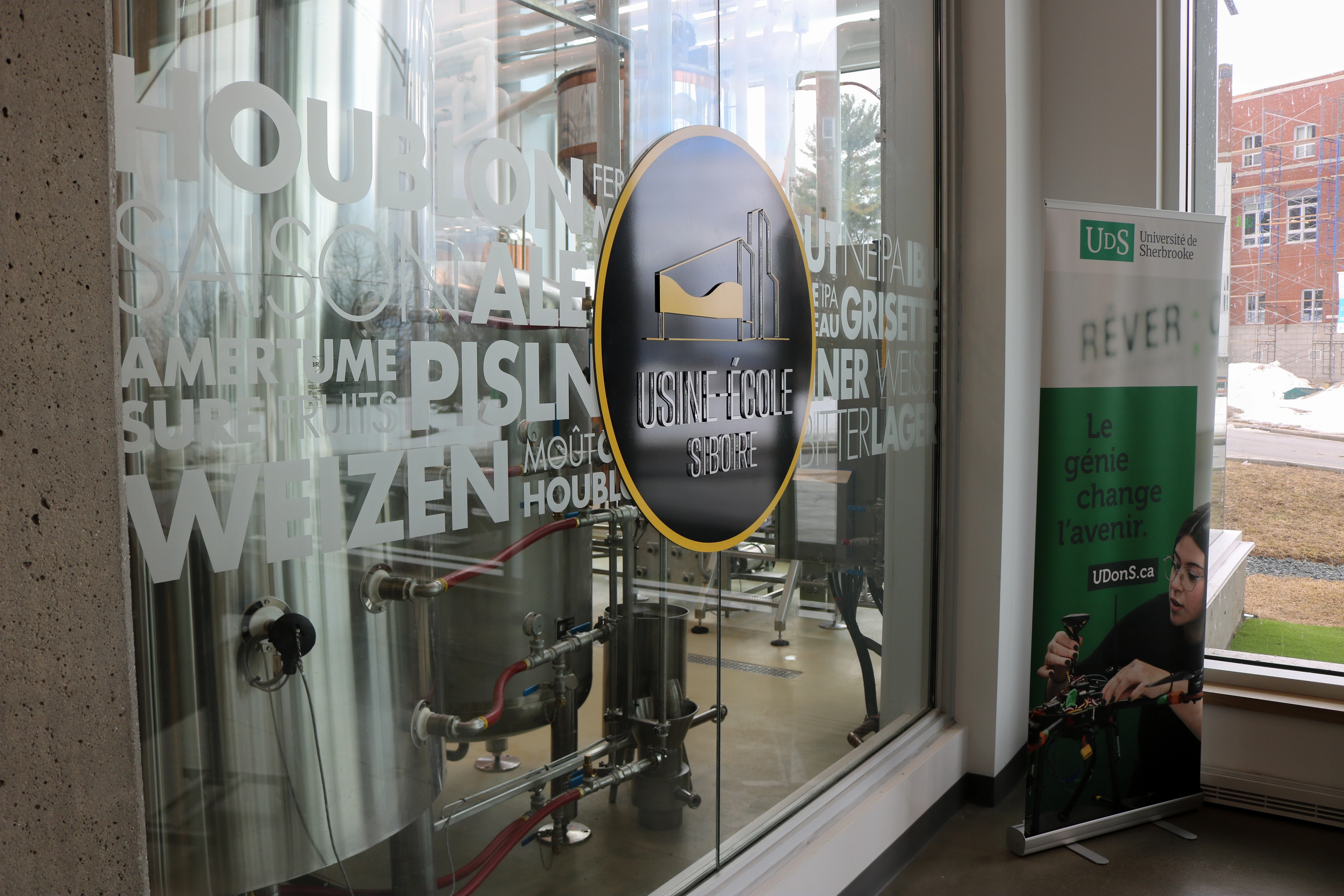 Glass wall of Usine-École Siboire brewery with industrial brewing equipment visible inside and a banner for Université de Sherbrooke reading 'Le génie change l'avenir' next to a window.