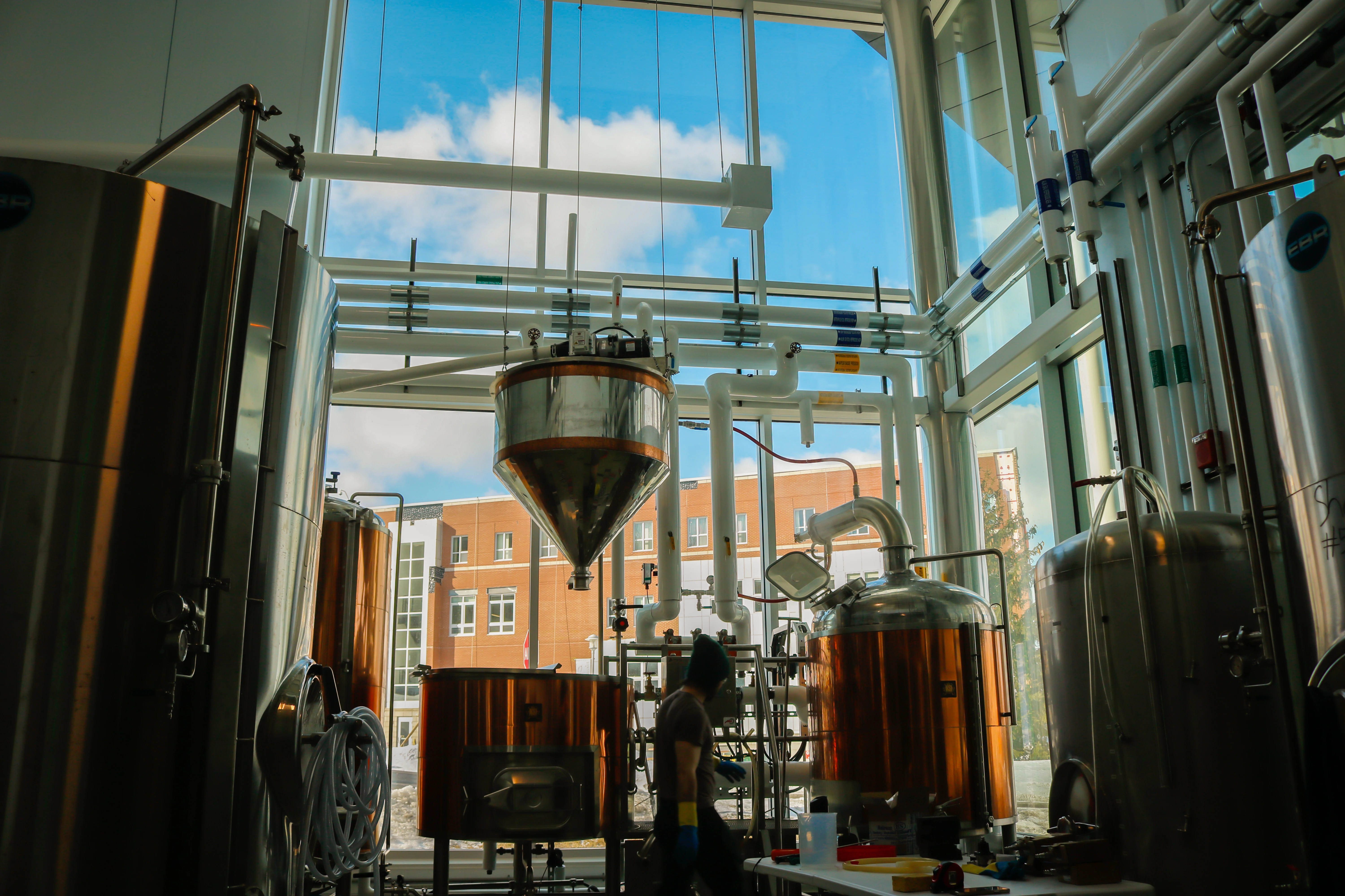 Interior of a brewery with large copper and stainless steel tanks, pipes, and a person walking by a table with brewing equipment.