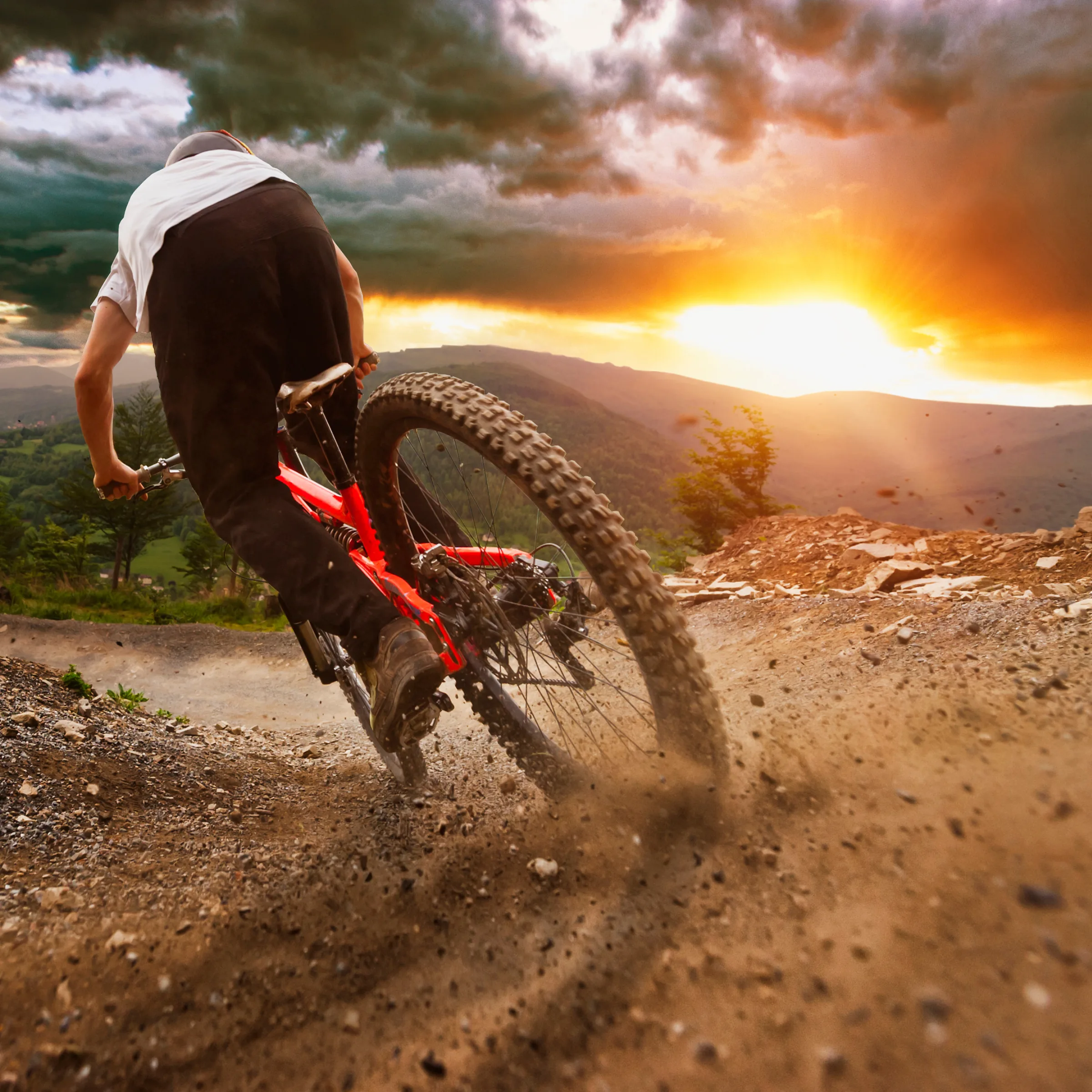 Person riding a red mountain bike fast down a dirt trail kicking up dust at sunset in hilly terrain.
