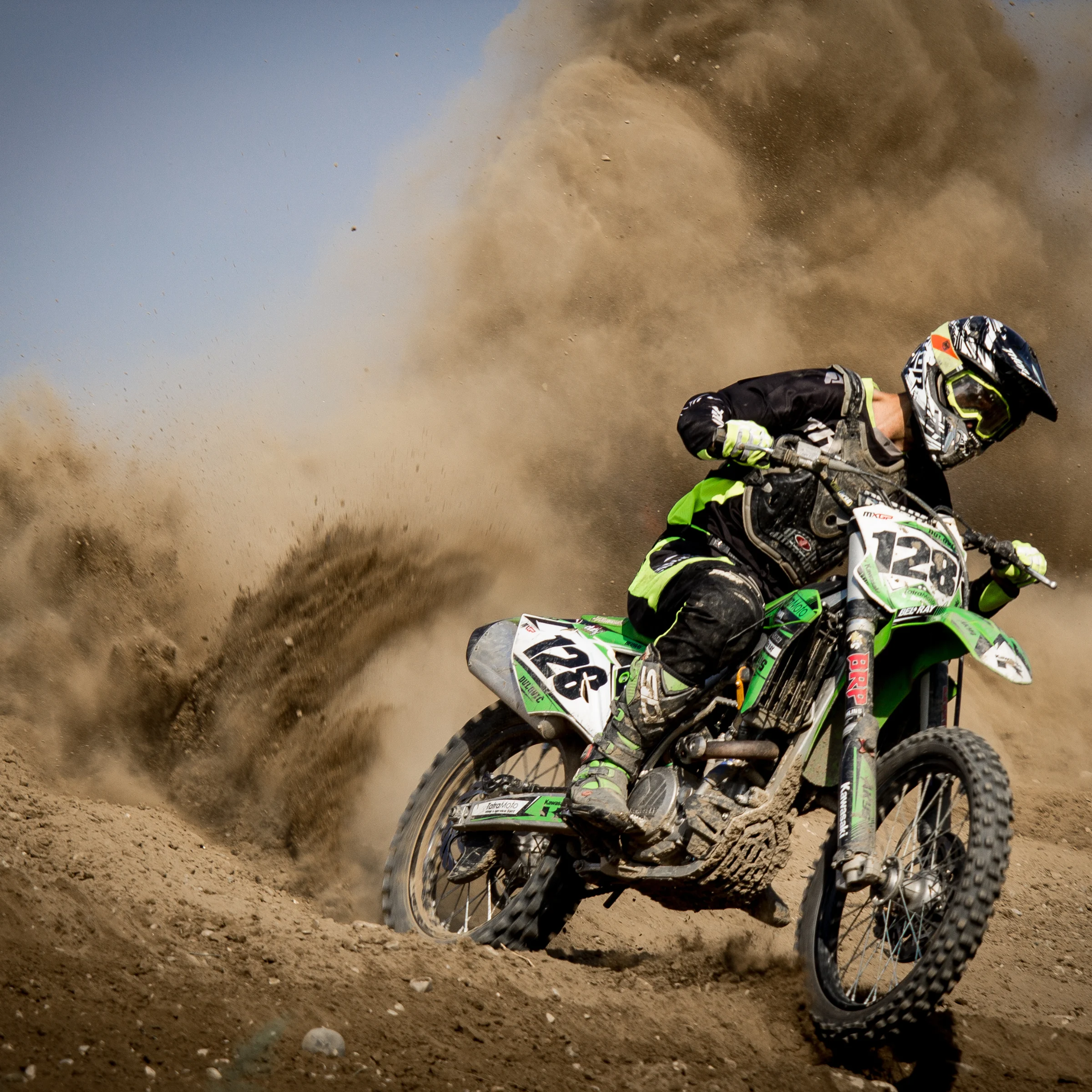 Motocross rider in black and green gear aggressively turning a dirt track, kicking up a large cloud of dust.