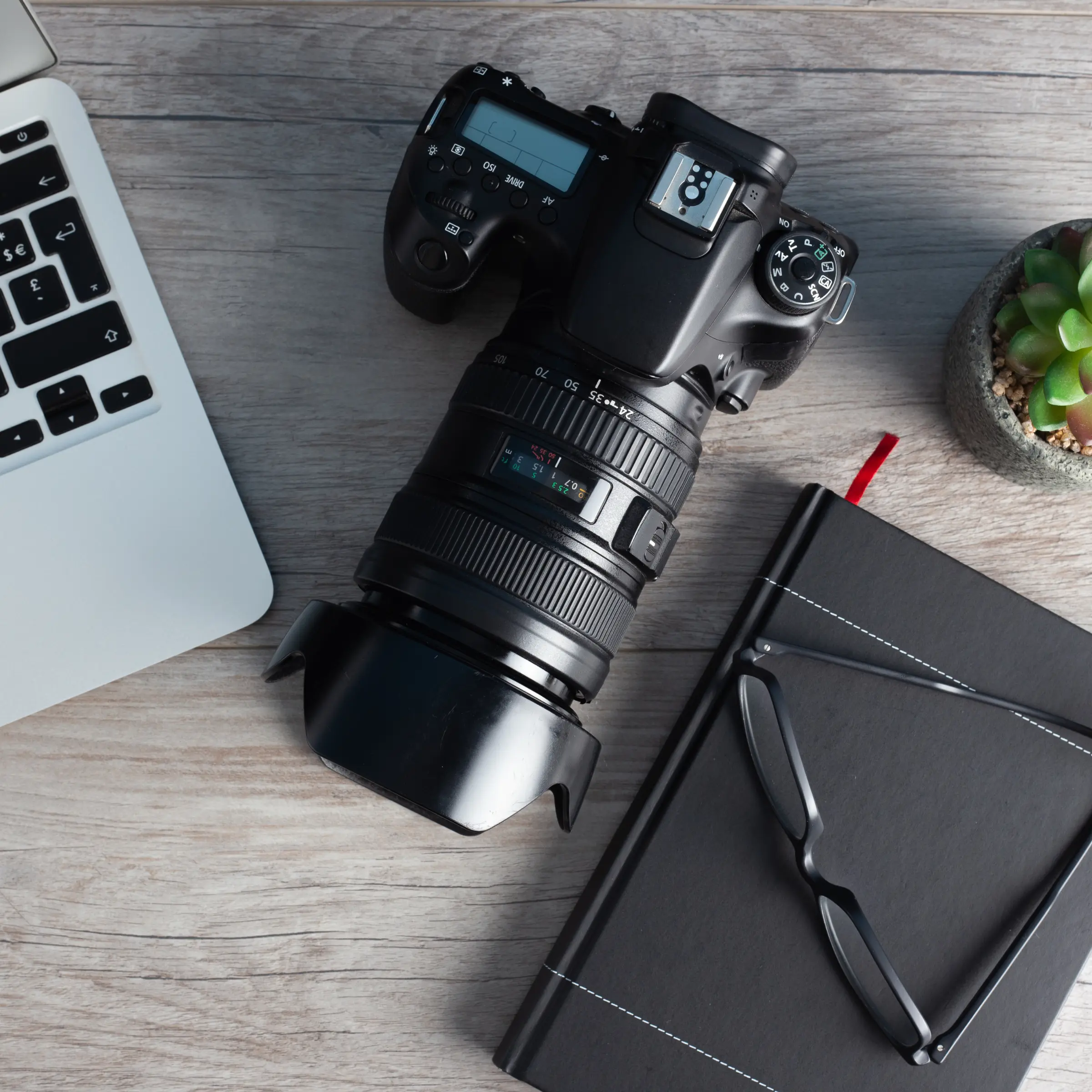 Black DSLR camera with lens and lens hood on a wooden desk, next to a laptop, black notebook with glasses, and a small succulent plant.