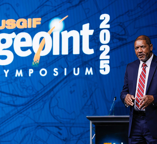 Man in suit and red-striped tie speaking on stage at the USGIF Geopoint 2025 Symposium with large event logo in background.
