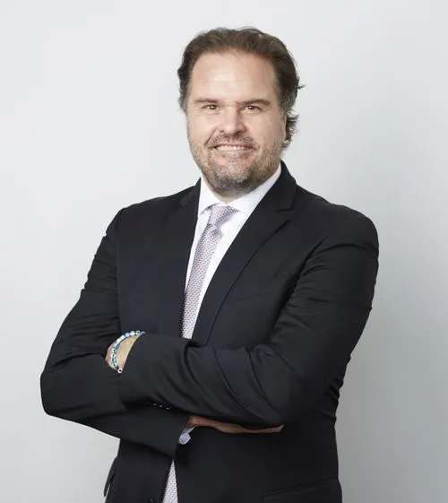 Smiling Chris Davis in a black suit with a light patterned tie, arms crossed, standing against a plain light gray background.