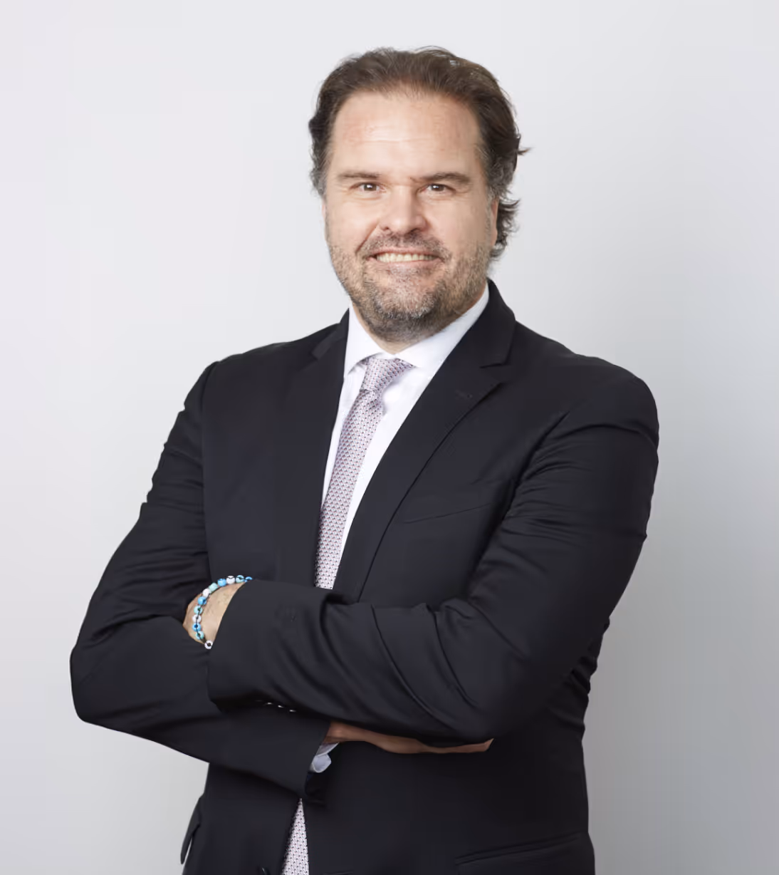 Smiling Chris Davis in a black suit with a light patterned tie, arms crossed, standing against a plain light gray background.