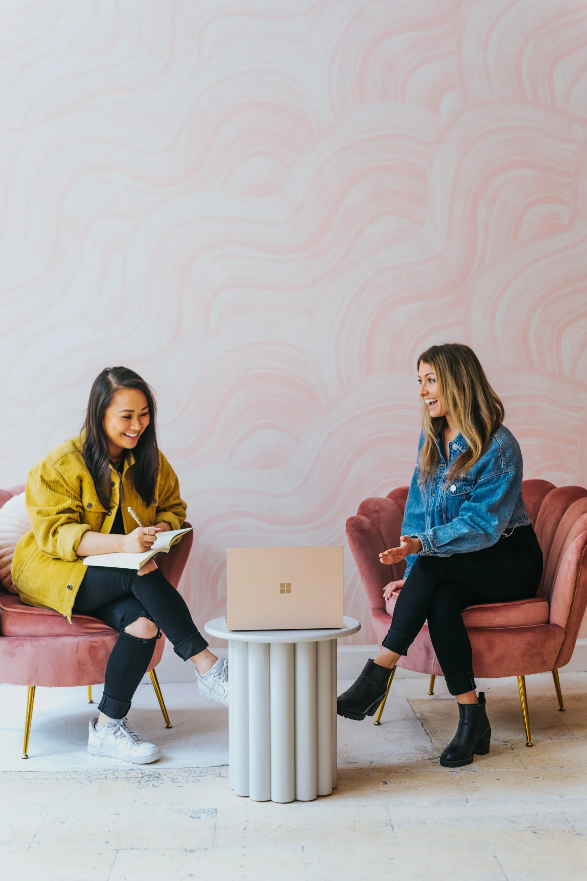 Two women sitting on pink chairs in front of a pink wall.