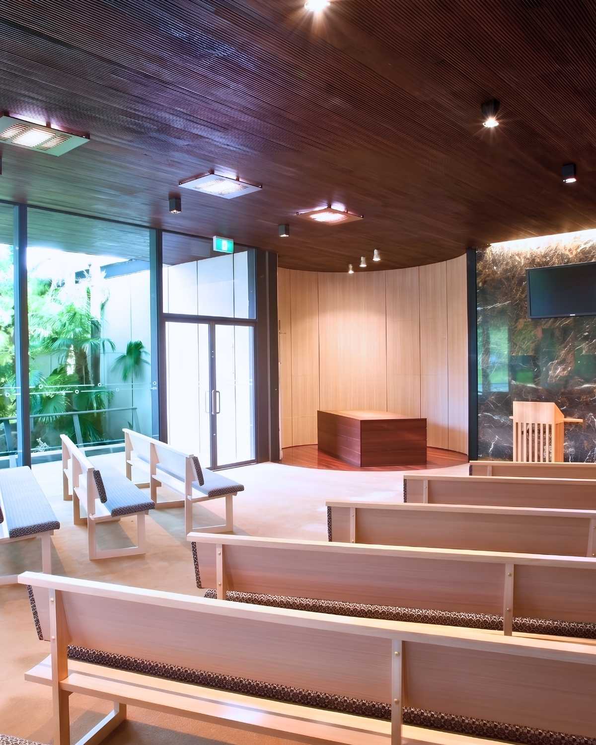 Quiet chapel interior with timber pews, curved panelling and a catafalque beneath a slatted ceiling