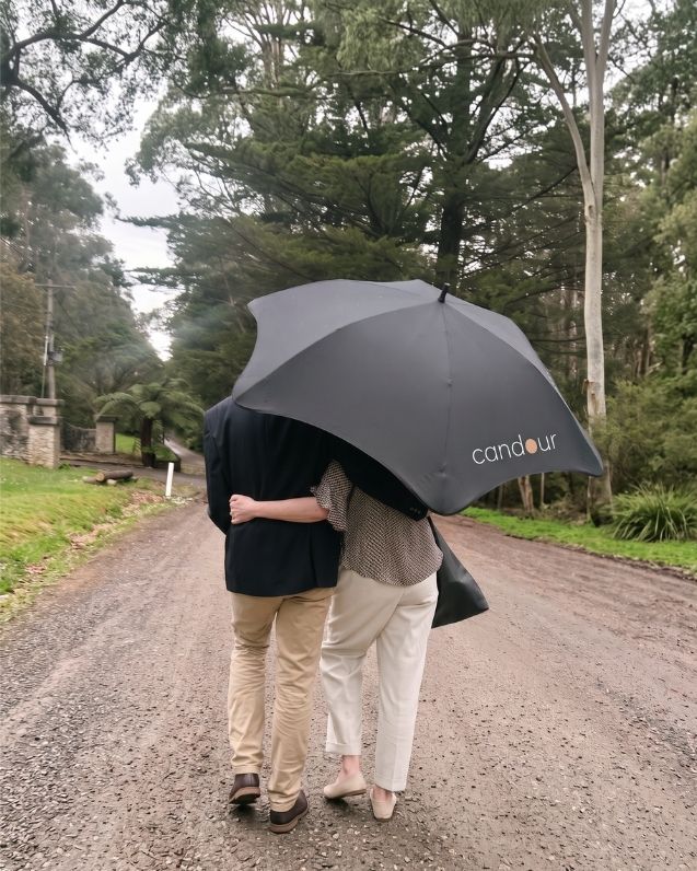 Two people walk arm in arm beneath a black Candour umbrella along a tree-lined gravel road.