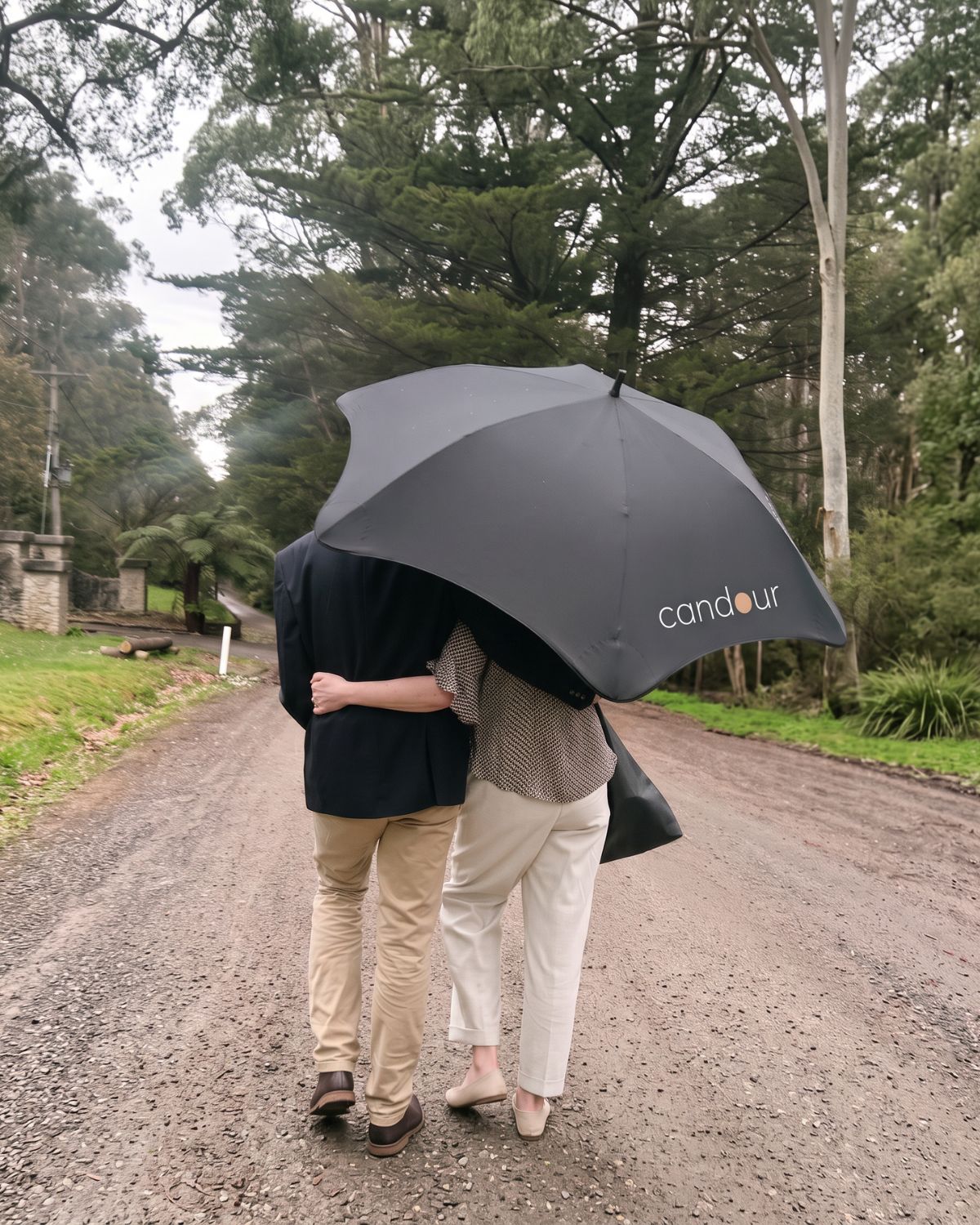 Two people walk arm in arm beneath a black Candour umbrella along a tree-lined gravel road.