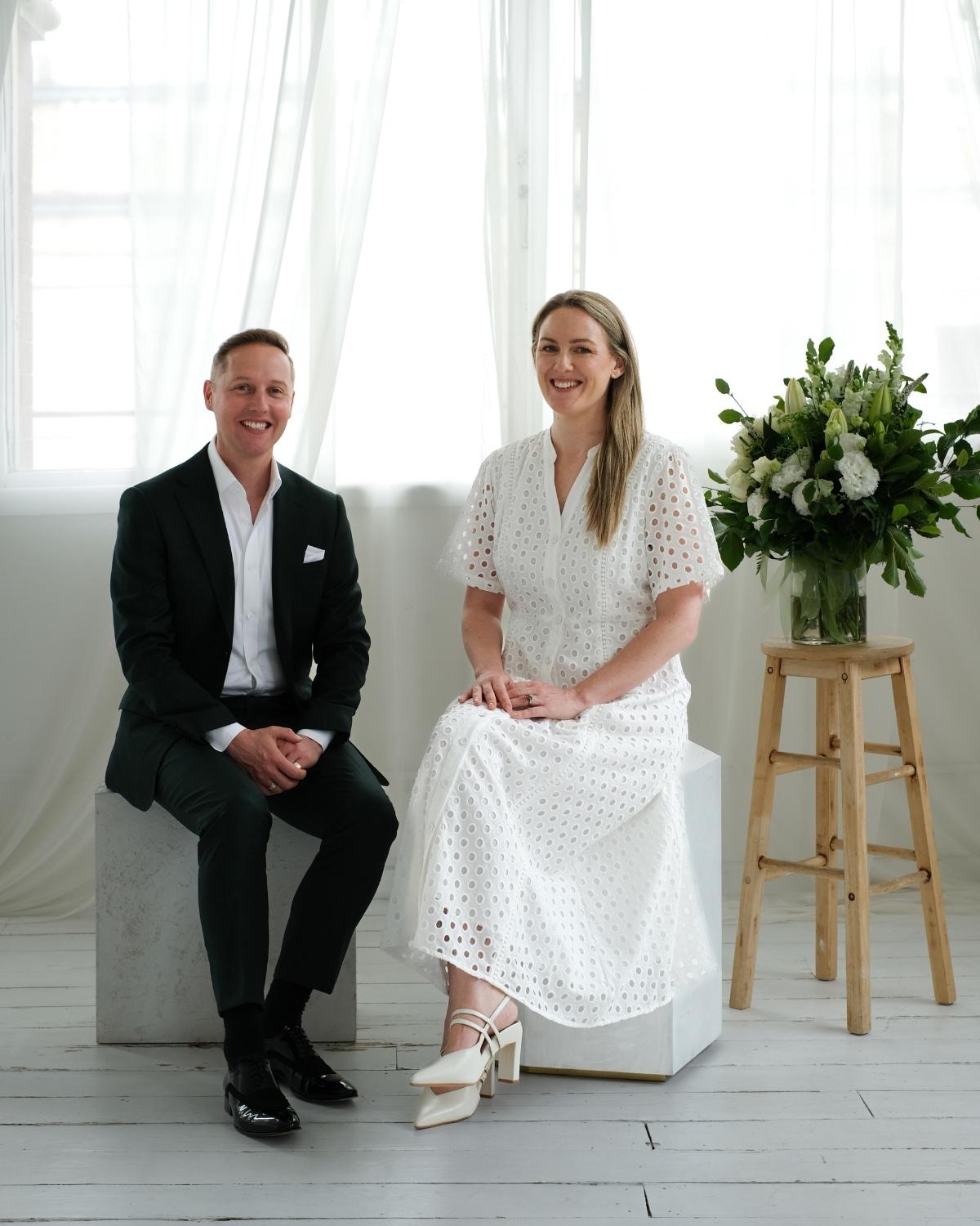 Simon Mumm&eacute; and Sameiko Slota, co-founders of Candour Funerals, seated together in a bright studio beside an arrangement of white flowers and native foliage.