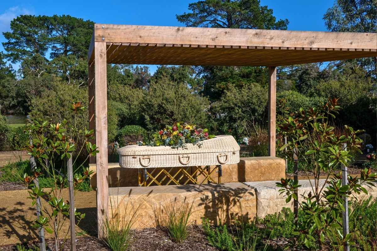 A white wicker coffin with native flowers resting under a timber pergola among established gardens