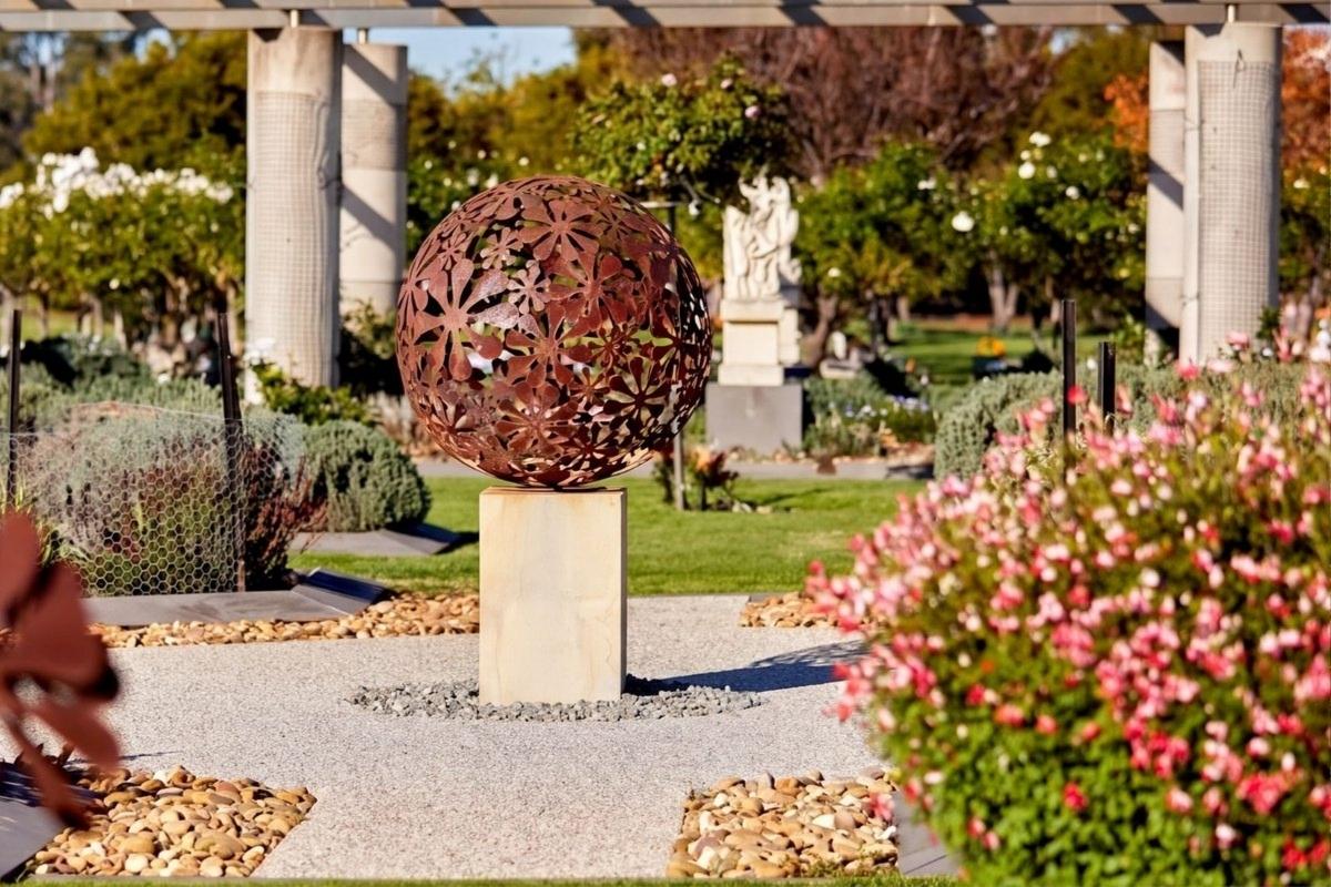A rusted steel sphere of cut flowers on a sandstone plinth among memorial garden beds in bloom