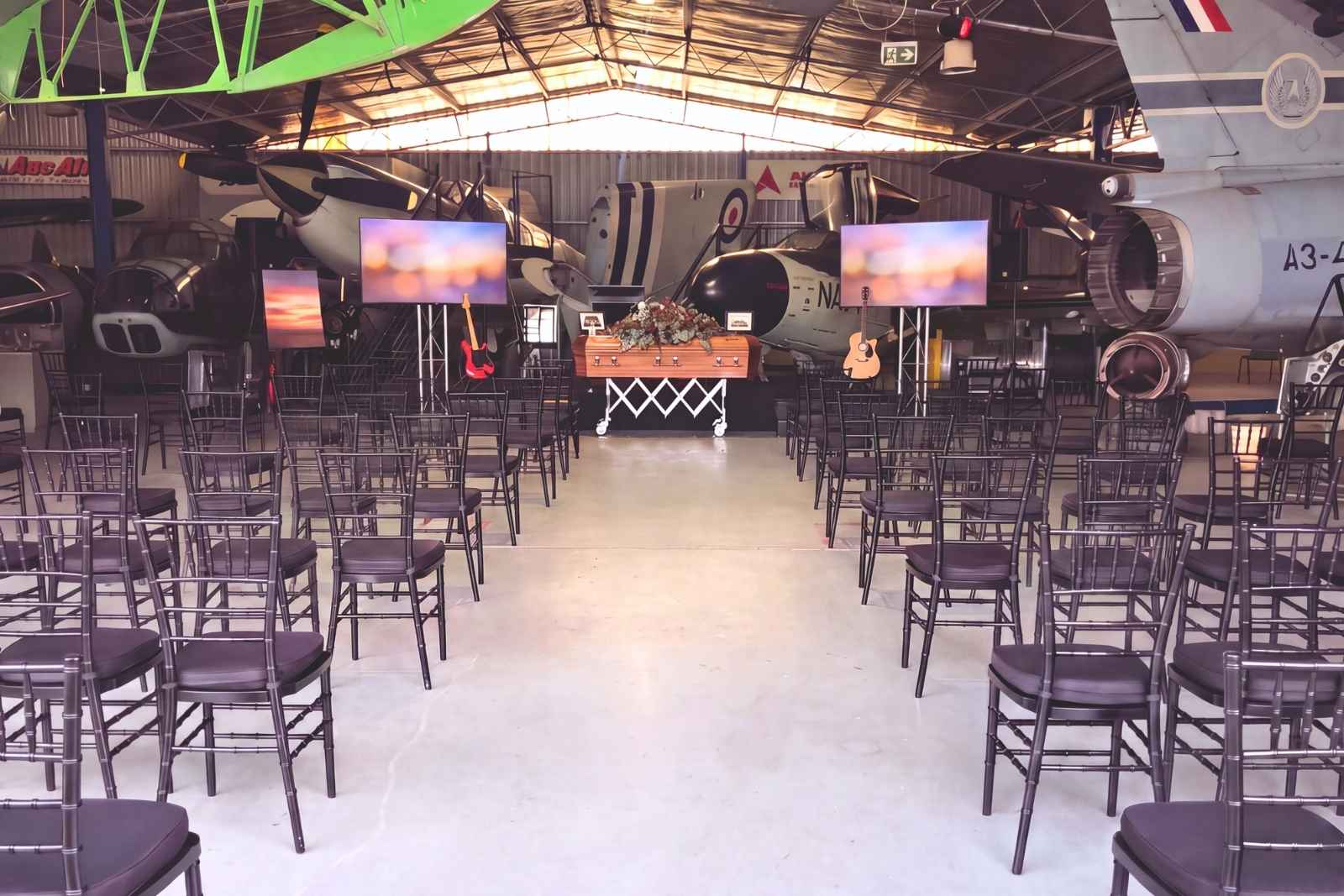 A farewell service set among aircraft at an aviation museum, chairs facing the coffin