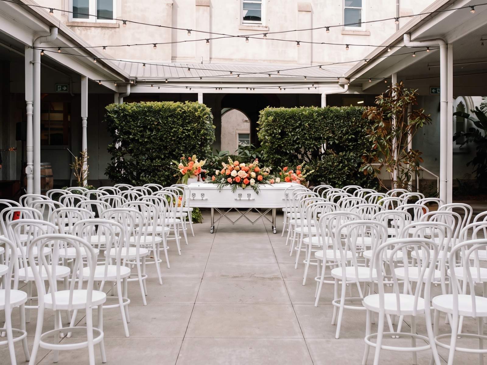 White chairs gathered for a courtyard farewell, a white coffin dressed with coral flowers