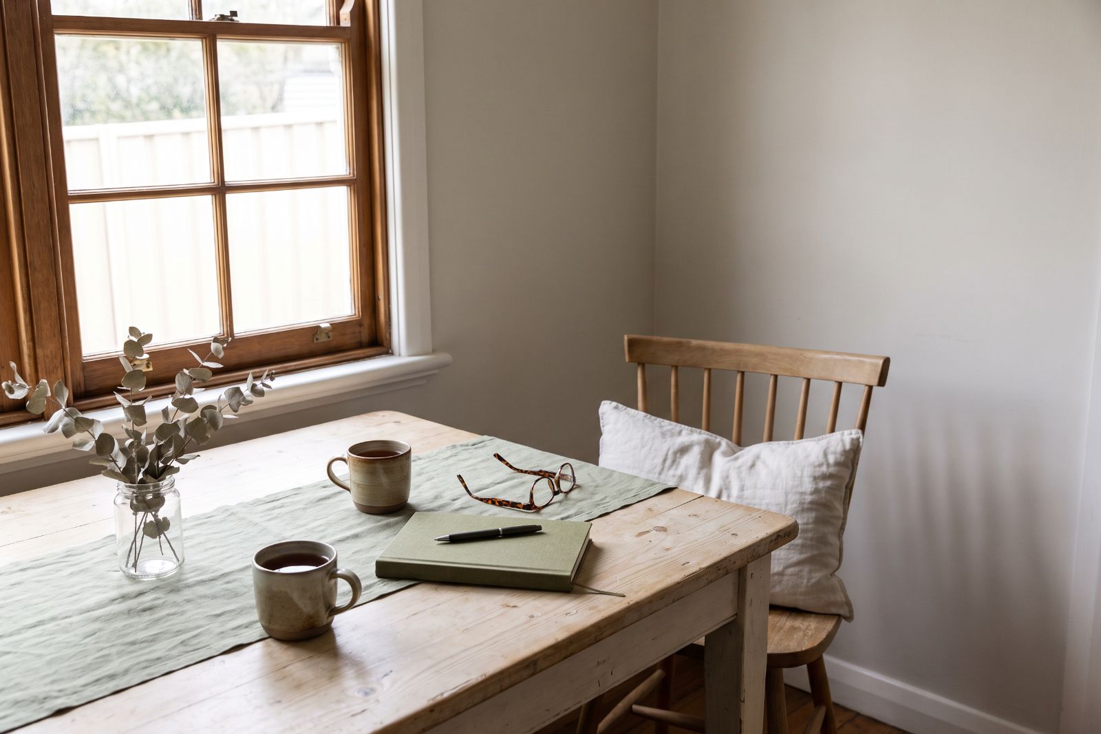 A kitchen table beside a window, with two cups of tea, a notebook and reading glasses