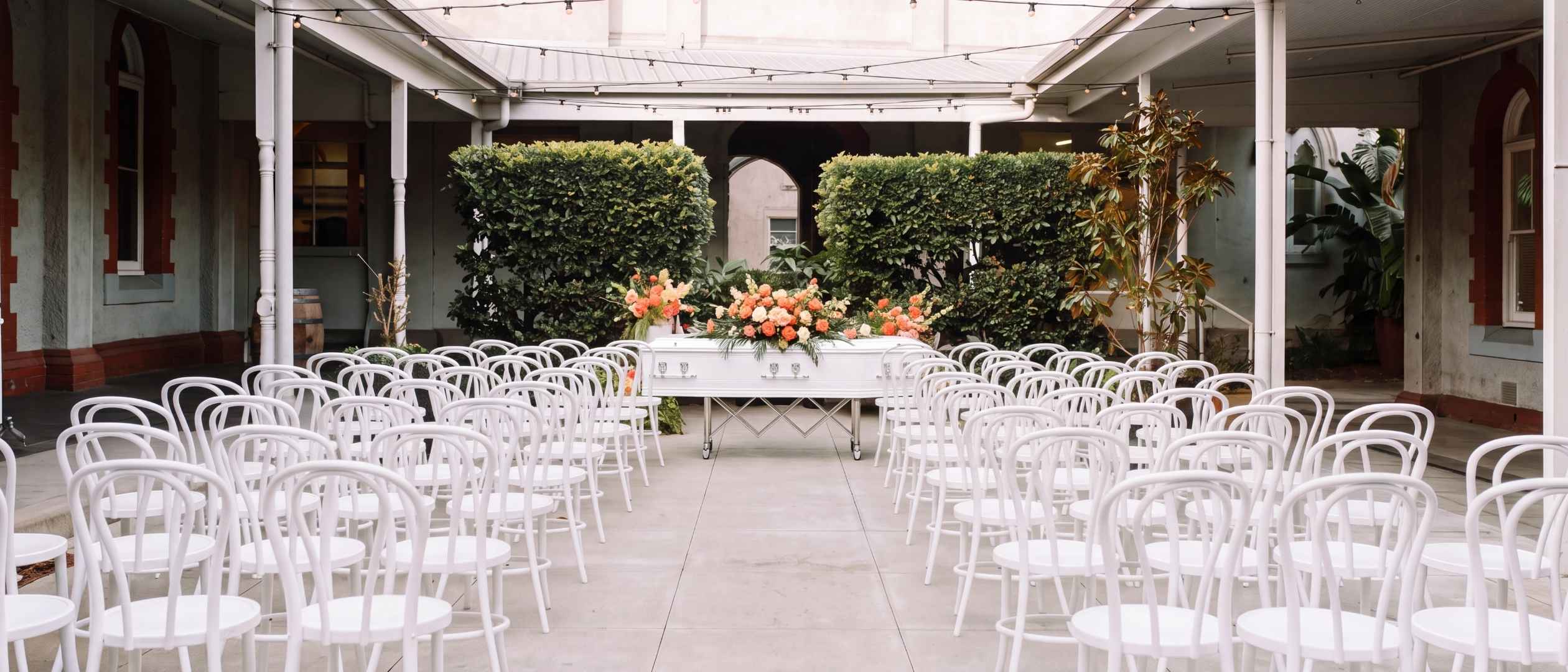 A courtyard ceremony set before guests arrive — white bentwood chairs, a white casket with coral florals beneath festoon lights