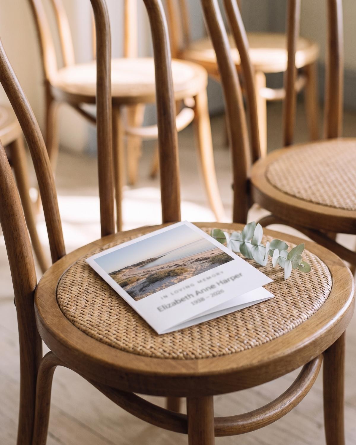 An order of service and a sprig of eucalyptus resting on a bentwood chair in soft light