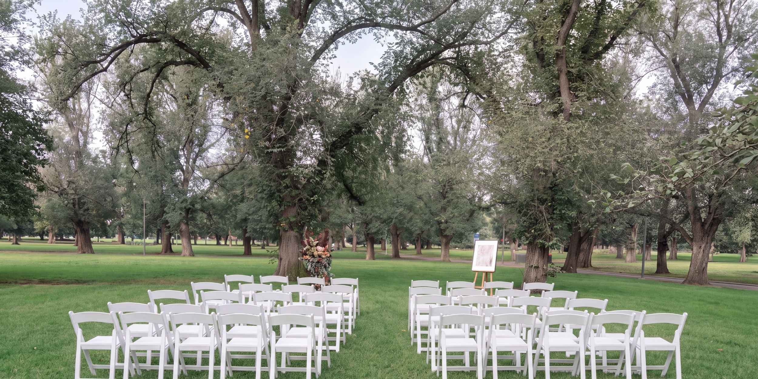 White chairs set on a lawn beneath old elms before a service, a floral tribute and portrait waiting