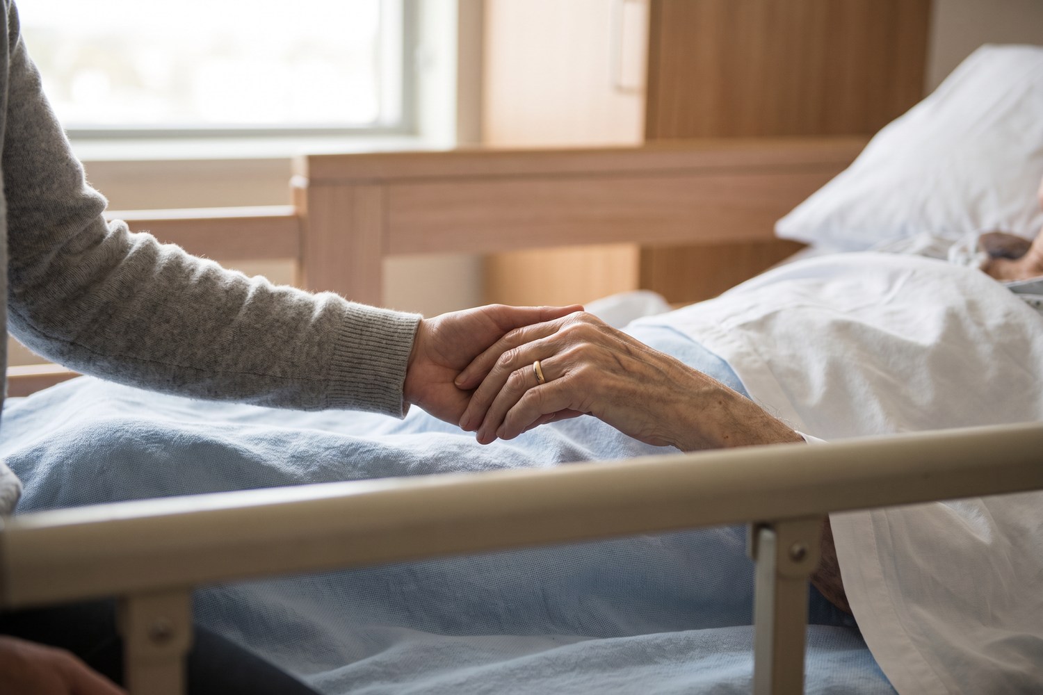 A family member holding an older person's hand beside a hospital bed