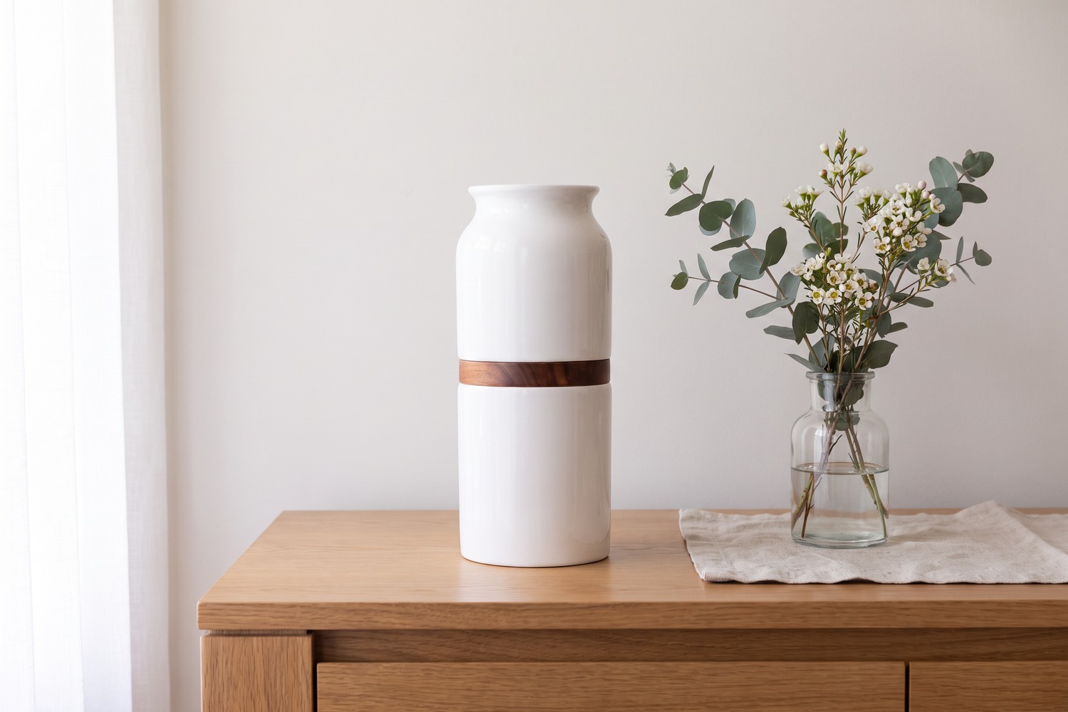 A plain ceramic urn with a timber band on a sideboard, beside a glass of eucalyptus and waxflower