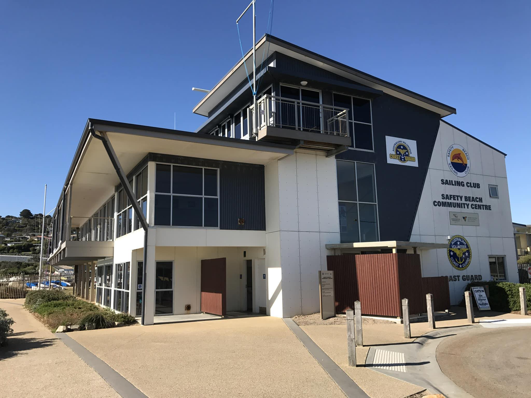 The exterior of the Safety Beach Sailing Club and Community Centre, a modern two-storey building with clean lines and nautical signage, located right by the coast under a clear blue sky.