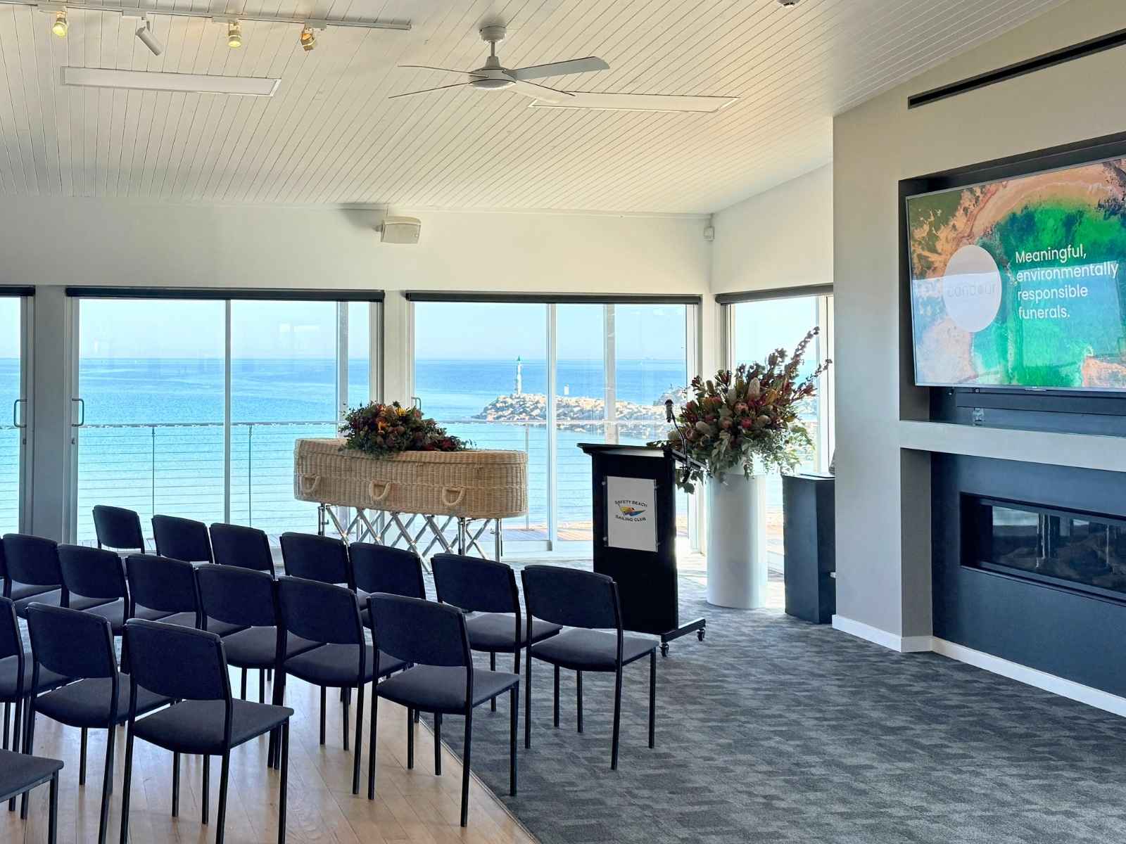 A funeral setup inside the Safety Beach Sailing Club, featuring rows of chairs, a natural woven coffin adorned with flowers, and large windows showcasing a panoramic ocean view.